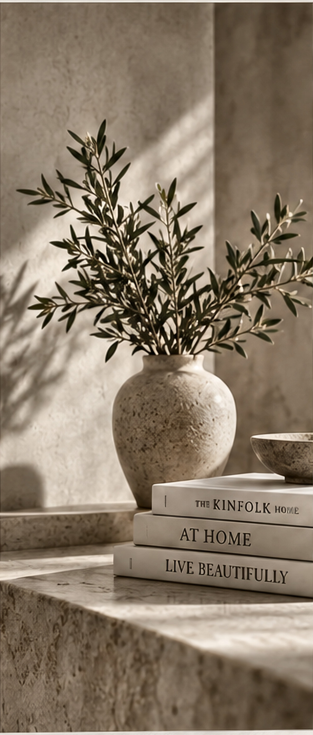 Olive branches in a stone vessel on a travertine plinth, beside a stack of interiors books
